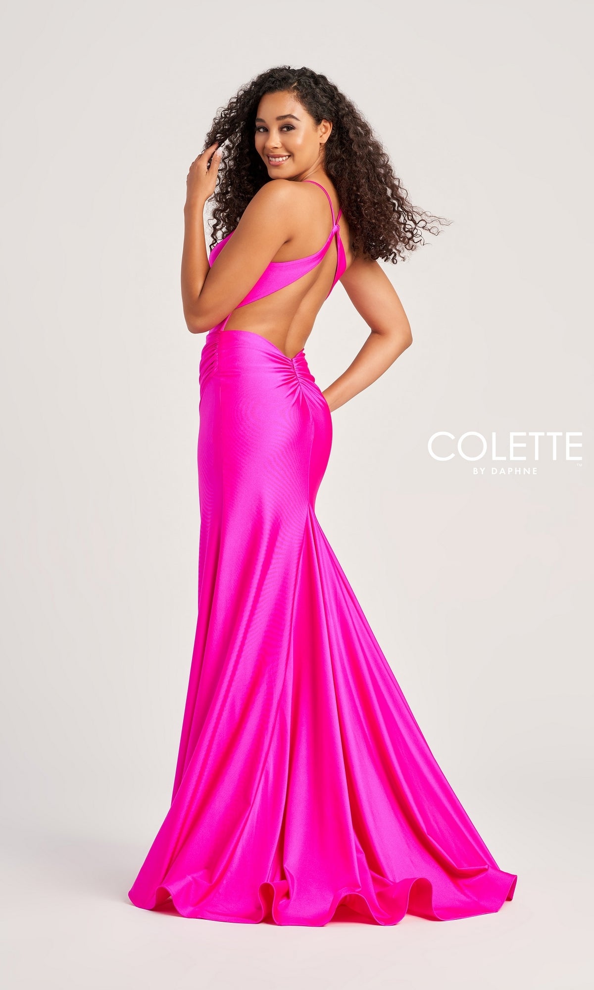 Woman in a bright pink, strappy-back, floor-length Sleek Long Colette Designer Prom Dress CL5204 with a fitted silhouette, posing sideways and smiling against a plain light background.